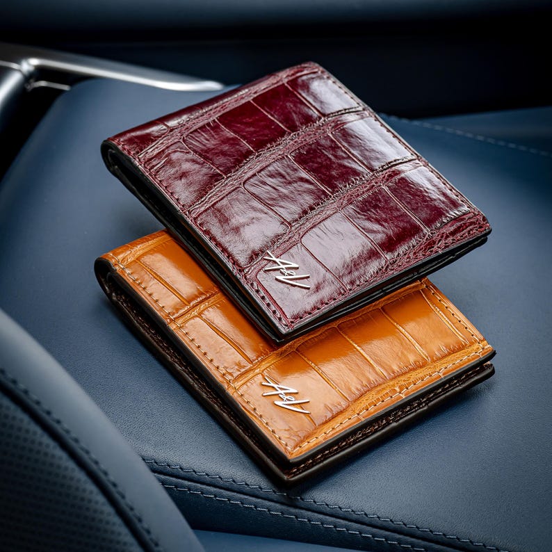 May include: Two leather wallets, one brown and one burgundy, with a crocodile skin pattern. Both wallets have the initials "A.L." embossed on the front.
