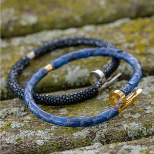May include: Two leather bracelets, one blue and one black, with gold accents. The bracelets are stacked on top of each other and are lying on a wooden surface.