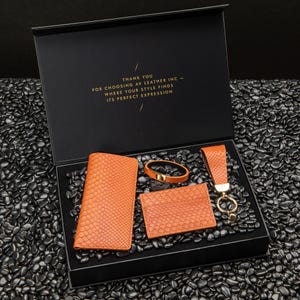 May include: A black gift box with a gold inscription that reads "THANK YOU FOR CHOOSING AV LEATHER INC - WHERE YOUR STYLE FINDS ITS PERFECT EXPRESSION". The box contains a set of orange leather goods, including a wallet, a bracelet, a card holder, and a key chain.