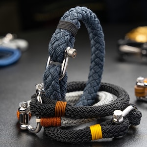 May include: A set of three braided rope bracelets with silver clasps. The bracelets are in shades of gray, black, and brown.