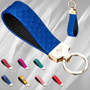 Może przedstawiać: Brązowy sk&oacute;rzany brelok do kluczy z złotym k&oacute;łkiem i złotą zawieszką z napisem "AV". Brelok wykonany jest ze sk&oacute;ry węża i ma czarne podłoże. W tle znajduje się dziewięć innych brelok&oacute;w w r&oacute;żnych kolorach.