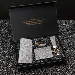 May include: A black gift box with a gray snakeskin wallet, a gray snakeskin card holder, a gray snakeskin bracelet, and a gold key chain. The box has a gold inscription that reads "Thank you for choosing AV Leather Inc - Where your style finds its perfect expression."