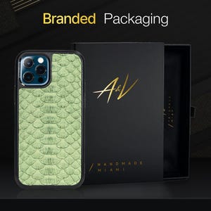 Pode incluir: Uma capa de telefone de pele de cobra verde em uma capa protetora preta. A capa é embalada em uma caixa preta com o logotipo "A&V HANDMADE MIAMI" impresso em dourado.
