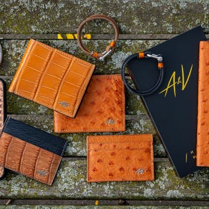 May include: Assortment of orange leather goods, including a phone case, wallets, and a passport holder. The items are arranged on a wooden surface with a black book with the letters "A&V" on the cover.