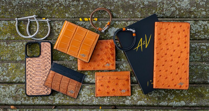 May include: Assortment of orange leather goods, including a phone case, wallets, and a passport holder. The items are arranged on a wooden surface with a black book with the letters "A&V" on the cover.