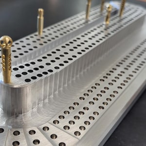 Può includere: Un tabellone di cribbage color argento con file di fori praticati per il posizionamento dei pioli. Il tabellone ha una forma rettangolare con angoli arrotondati e una serie di fori lungo i bordi per il punteggio. I pioli sono di colore oro.