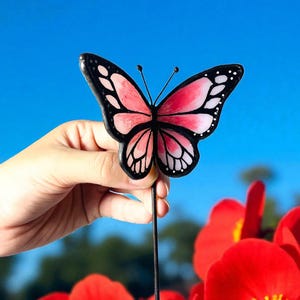 May include: A garden ornament featuring a butterfly with pink and white wings, edged in black, held against a blue sky. The butterfly has a black body and antennae. Red flowers with yellow centres are in the foreground.