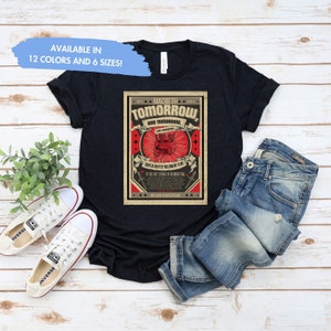 May include: A black t-shirt with a vintage-style graphic design featuring the text "MACBETH TOMORROW AND TOMORROW" in red and white. The graphic also includes a skull and crossbones with a red heart. The text "IN THE LAST SYLLABLE OF RECORDED TIME" is also included in the graphic.