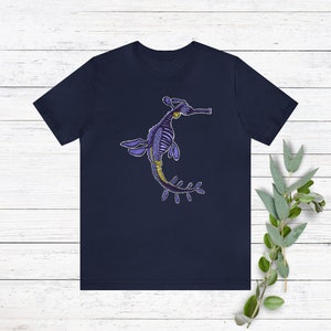 K&ouml;nnte beinhalten: Ein marineblaues T-Shirt mit einem Aufdruck eines lila und gelben Seepferdchens mit blattf&ouml;rmigen Anh&auml;ngseln.