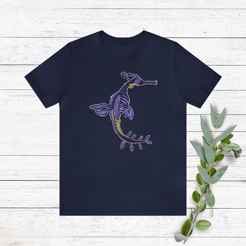 K&ouml;nnte beinhalten: Ein marineblaues T-Shirt mit einem Aufdruck eines lila und gelben Seepferdchens mit blattf&ouml;rmigen Anh&auml;ngseln.