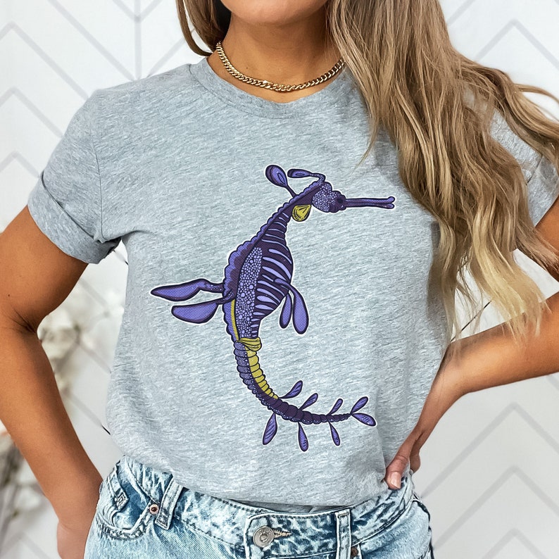 K&ouml;nnte beinhalten: Ein graues T-Shirt mit einem lila und gelben Seepferdchen-Design. Das Seepferdchen ist mit einer Skelettstruktur detailliert und hat einen langen, flie&szlig;enden Schwanz.