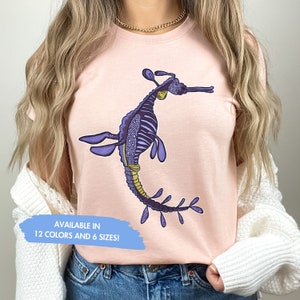 K&ouml;nnte beinhalten: Ein pinkfarbenes T-Shirt mit einer Illustration eines lila und gelben Seepferdchens. Der Text "AVAILABLE IN 12 COLORS AND 6 SIZES!" befindet sich auf einem blauen Hintergrund.