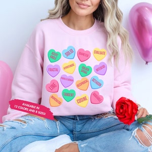 May include: A pink sweatshirt with a graphic of colorful hearts featuring math terms like 'acute angle', 'solid', 'diameter', 'evaluate', 'fractal', 'hypotenuse', 'golden spiral', 'integers', 'line', 'parallelogram', 'reciprocal', 'octagon', 'numerator', 'tangent', 'cosine', and 'variable'. The text 'AVAILABLE IN 12 COLORS AND 8 SIZES!' is at the bottom of the sweatshirt.