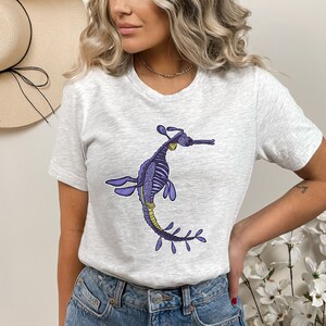 K&ouml;nnte beinhalten: Ein graues T-Shirt mit einer Illustration eines lila und gelben Seepferdchens. Das Seepferdchen hat einen langen, d&uuml;nnen Schwanz und einen kleinen Kopf mit einer langen Schnauze.