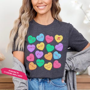 Puede incluir: Camiseta gris oscuro con caramelos en forma de corazón coloridos con ecuaciones y símbolos matemáticos. El texto "AVAILABLE IN 12 COLORS AND 6 SIZES!" está impreso en la parte inferior izquierda. La camiseta es de estilo casual.