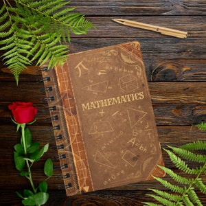 May include: A brown leather-bound notebook with a spiral binding. The cover features the word "MATHEMATICS" in gold lettering and various mathematical formulas and diagrams. The notebook is lying on a wooden surface with a green fern and a red rose.