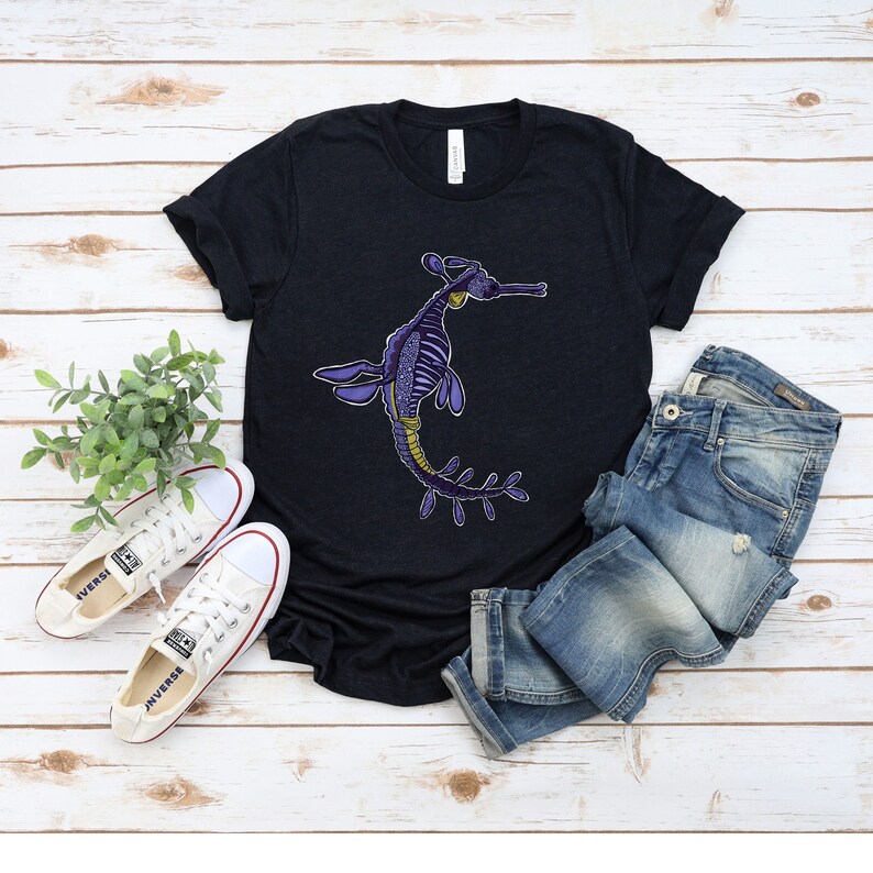 K&ouml;nnte beinhalten: Ein schwarzes T-Shirt mit einer Illustration eines lila und gelben Seepferdchens. Das T-Shirt liegt auf einer wei&szlig;en Holzoberfl&auml;che mit einem Paar wei&szlig;en Sneakers und einer blauen Jeans.
