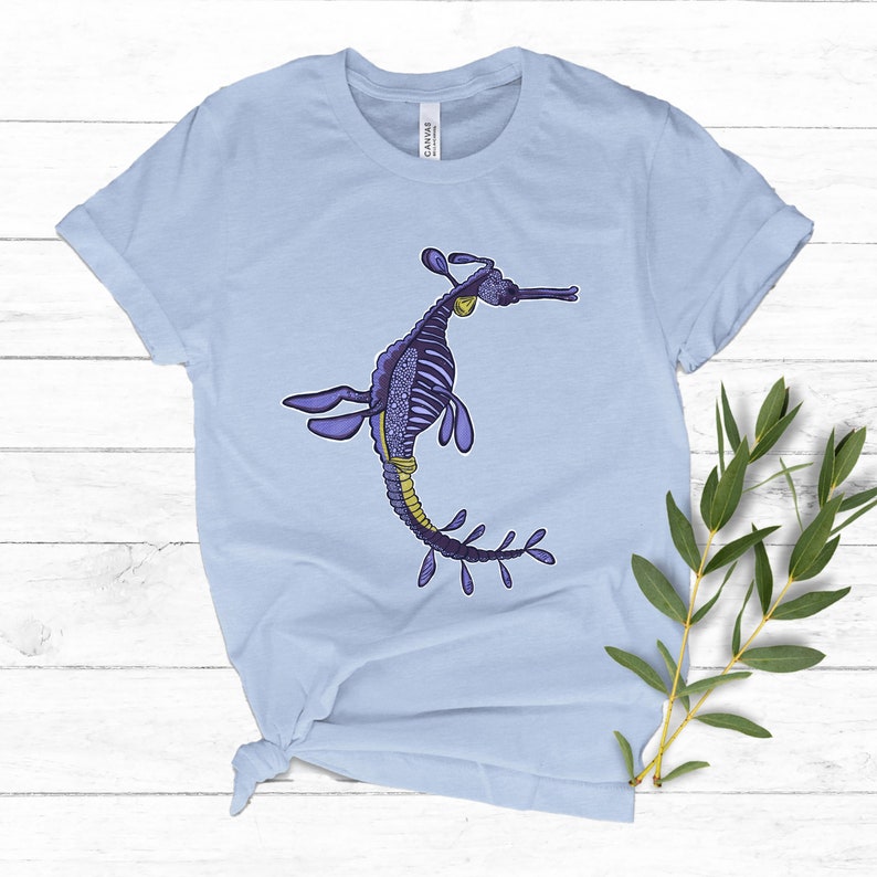 K&ouml;nnte beinhalten: Hellblaues T-Shirt mit einer Illustration eines lila und gelben Seepferdchens. Das Seepferdchen ist mit einem Skelettmuster verziert und hat einen langen, flie&szlig;enden Schwanz.