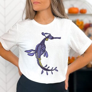 K&ouml;nnte beinhalten: Wei&szlig;es T-Shirt mit einer Illustration eines lila und goldenen Seepferdchens. Das Seepferdchen ist mit einem Skelettdesign verziert und hat einen langen, flie&szlig;enden Schwanz.