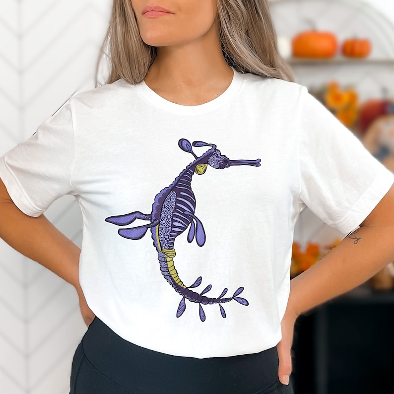 K&ouml;nnte beinhalten: Wei&szlig;es T-Shirt mit einer Illustration eines lila und goldenen Seepferdchens. Das Seepferdchen ist mit einem Skelettdesign verziert und hat einen langen, flie&szlig;enden Schwanz.