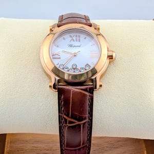 May include: A rose gold wristwatch with a white face, Roman numeral hour markers, and a brown leather band. The watch face features the brand name "Chopard" and small diamonds. The band is textured and appears to be made of leather.