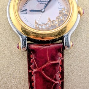 May include: A luxury Chopard Geneve watch with a gold and silver case, a mother-of-pearl face, and a burgundy leather strap. The watch face features a moon and star design with diamonds. The strap is approximately 7 inches long.