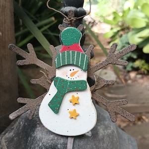 May include: A wooden snowman ornament with brown twig arms, a green and red striped hat, a green scarf with red polka dots, and two yellow stars.