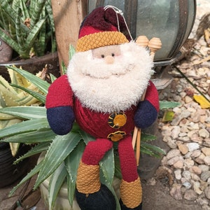 May include: A plush Santa Claus doll with a red and white suit, a white beard, and a brown knit hat. The doll is sitting on a green plant with brown stems.