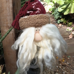 May include: A white-bearded gnome with a red and brown hat. The gnome is made of fabric and has a round, pink nose.