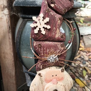 May include: A wooden Christmas ornament shaped like a Santa Claus with a red hat, white beard, and brown eyes. The ornament is decorated with a snowflake and a wreath of twigs and berries.