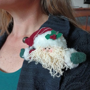 May include: A small, white, plush Santa Claus ornament with a red hat, green holly, and a white beard. The ornament is attached to a dark gray sweater.