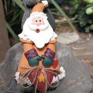 May include: A ceramic Christmas ornament featuring a Santa Claus figure with a red nose and a white beard. He is holding a red heart with a gold star in the center, tied with twine.