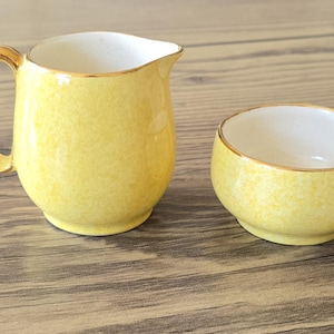 May include: A yellow ceramic creamer jug with a handle and a matching small open sugar bowl. Both pieces feature a mottled yellow exterior finish, a smooth white interior, and a thin gold trim along the top rims and the jug handle.