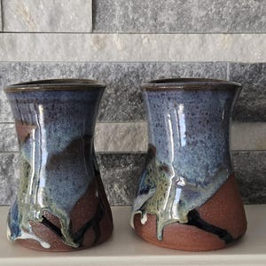 May include: Two ceramic tumblers with a flared rim and a tapered base. The surface features a mottled blue and grey glaze that transitions into a dark brown, unglazed clay bottom, with visible drip patterns where the glaze meets the raw clay.