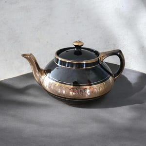 May include: A black ceramic teapot featuring a glossy finish, a matching lid with a small knob, and a curved handle. The body is decorated with a wide, metallic gold band showcasing an intricate, repeating scalloped pattern around the centre.