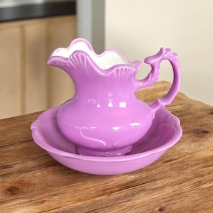 May include: A lavender ceramic washbasin set consisting of a pitcher with a scalloped rim and a matching round bowl. The pitcher features a curved handle and a white interior, while the bowl has a smooth, wide edge and a solid lavender finish.