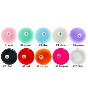 Op de afbeelding: Tien ronde plastic schijven in verschillende kleuren, waaronder roze, groen, blauw, wit, paars, zwart, rood, oranje, felroze en transparant. Elke schijf heeft een klein gaatje in het midden.