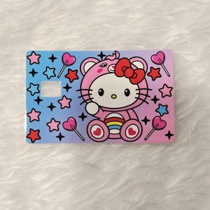 May include: A rectangular card featuring a pink Hello Kitty character in a pink outfit with a red bow. The card has a gradient background of blue and pink, decorated with stars and heart-shaped lollipops.
