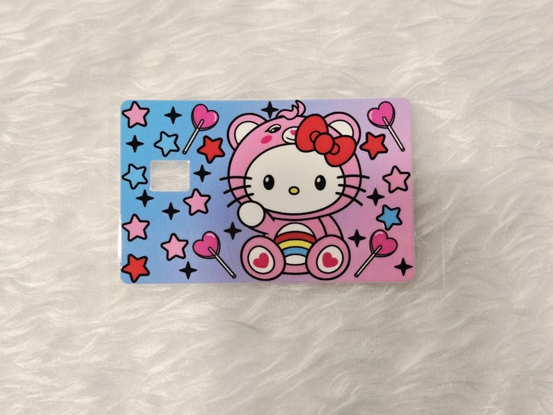 May include: A rectangular card featuring a pink Hello Kitty character in a pink outfit with a red bow. The card has a gradient background of blue and pink, decorated with stars and heart-shaped lollipops.
