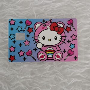 May include: A rectangular card featuring a Hello Kitty design in a pink bear costume with a red bow. The card has a gradient background of blue and pink, decorated with stars and heart-shaped lollipops. There is a square cut-out on the card.