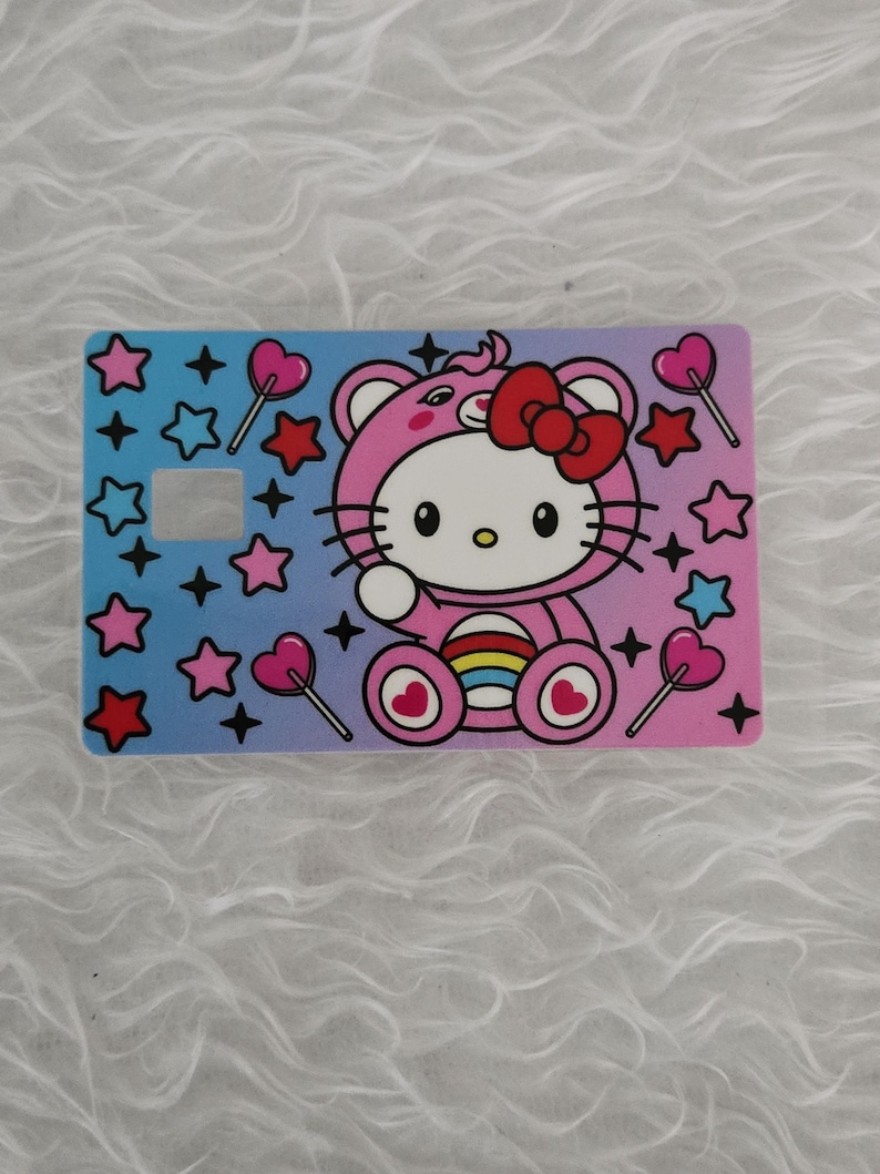 May include: A rectangular card featuring a Hello Kitty design in a pink bear costume with a red bow. The card has a gradient background of blue and pink, decorated with stars and heart-shaped lollipops. There is a square cut-out on the card.