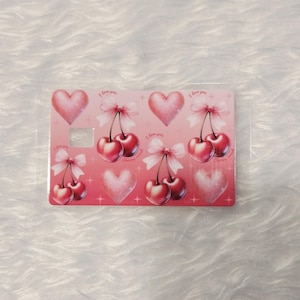 May include: A pink card with a pattern of cherries, hearts, and bows. The cherries are red with green stems and white bows. The hearts are pink, and the text "I love you" is visible. The card is in a clear plastic sleeve.