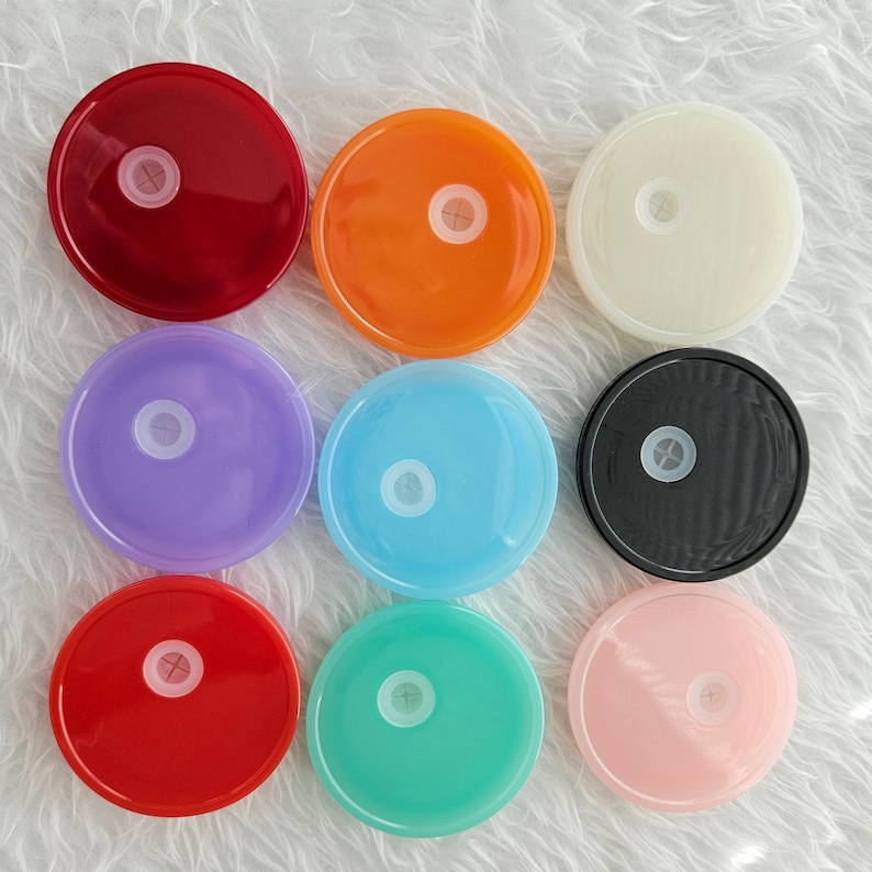 Op de afbeelding: Negen ronde deksels in verschillende kleuren, waaronder rood, oranje, wit, paars, lichtblauw, zwart, roze en turquoise. De deksels zijn allemaal even groot en hebben een klein gaatje in het midden.