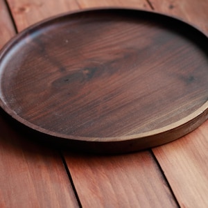 May include: A round, dark brown wooden tray with a raised edge. The tray is made of a rich, dark wood and sits on a wooden surface. The tray is approximately 12 inches in diameter.