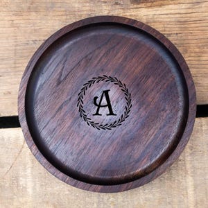 May include: A round, dark brown wooden tray with a raised edge. The tray features a black laurel wreath design encircling the letter "A" in the center. The wood grain is visible, adding texture to the surface.