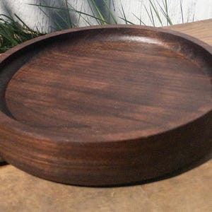 May include: A round, dark brown wooden tray with a raised edge. The tray has a smooth, polished surface and appears to be made of a solid piece of wood. The tray is sitting on a wooden surface.