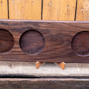May include: A dark brown wooden serving tray with four circular indentations for glasses. The tray has a handle on one end and is displayed on a wooden surface. The wood grain is visible.
