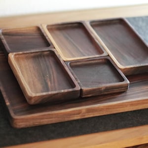 May include: A set of wooden trays in various sizes, arranged in a nested configuration. The trays are made of dark-stained wood, showcasing the natural grain. The set includes a large rectangular tray and several smaller, square and rectangular trays.
