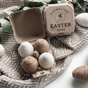 May include: A cardboard egg carton with the words "Farmhouse Easter Eggs" printed on the lid. The carton contains four decorative eggs wrapped in twine, two brown and two white.