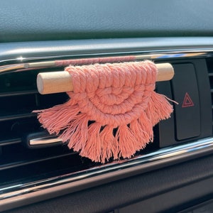 May include: A pink macrame car air freshener with tassels hanging from the bottom. The air freshener is attached to a wooden stick and is hanging from a car's air vent.